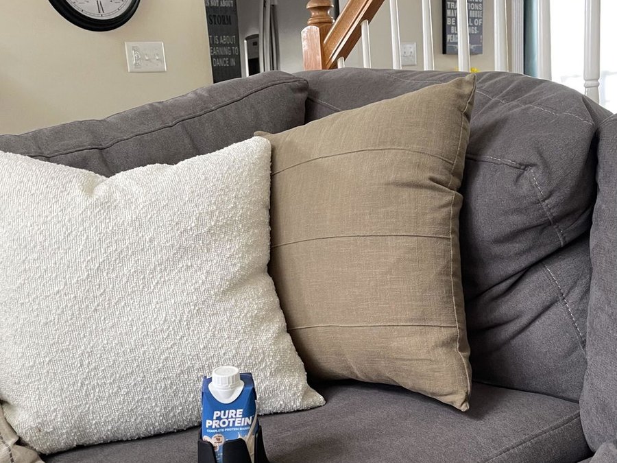 A black drink base holding a Pure Protein shake sitting on the seat cushion of a grey sectional sofa between a white boucle pillow and a tan pillow; wall clocks, staircase and kitchen behind
