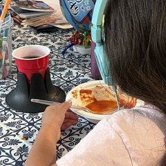 A young girl in mint headphones eating a plate of food at a kitchen table with a blue-and-white tiled tablecloth; a black drink base holds a red plastic party cup beside her plate, a birthday balloon behind