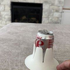 A Diet Dr Pepper can in a white drink base standing on a soft taupe blanket, a hand steadying nothing beside it, with a stone fireplace and a wall-mounted TV in the background