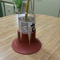 A clear glass drink bottle with a yellow straw sitting in a brick-red drink base on a light oak kitchen table, pale green cabinets and a kitchen counter behind